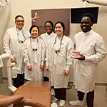 Five people in lab coats standing behind a dental chair. One of the people is holding a model of top and lower jaws with teeth.