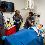 Four people in scrubs standing in a hospital room. An adult mannequin is laying on the bed. One person has their hands on the mannequin torso.