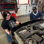 Two people in an auto body shop kneeling in front of a car with the front hood taken off.