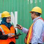 Two people wearing orange vests and hard hats standing in front of a green wall.