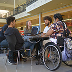 four people sitting around a table. One of the people has a laptop open in front of them. One of the people is sitting in a wheelchair.