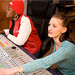 Two people sitting at a mixing board in a music recording studio. One of the people has a red knit hat on. The other person has one of their hands on the mixing board.