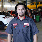 Person with mechanics shirt on standing in a car service shop with several cars behind them.