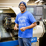 Person with blue knit cap and United States Air Force t-shirt on standing in a manufacturing shop in front of a machine.