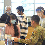 Four people in a lab. Two of the people are wearing masks over their mouths and noses. The third person is wearing a mask over their nose and mouth as well as a face shield. The fourth person is wearing a military uniform.
